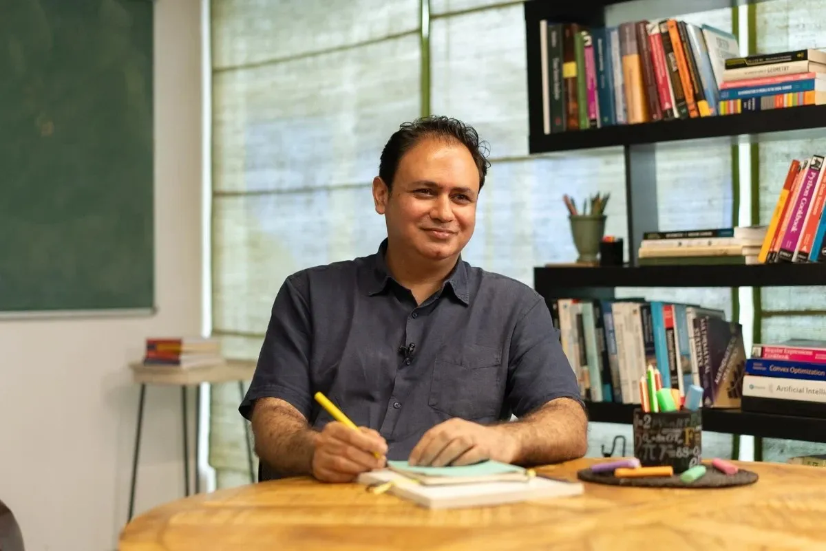 A smiling man sitting at a wooden table, writing in a notebook with a yellow pencil, surrounded by books and stationery in a well-lit room.